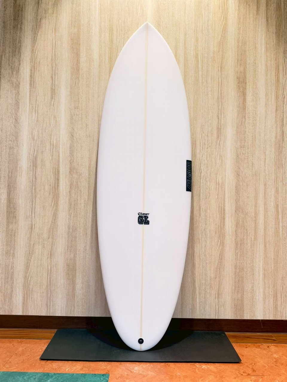 LINE_ALBUM_2026 フリーダム_260601_1 - SHOP NEWS | ムラサキスポーツオンラインストア 通販 White surfboard standing upright on a black mat against a wood-patterned wall, showing a small logo near the center and a vertical tag on the right edge.