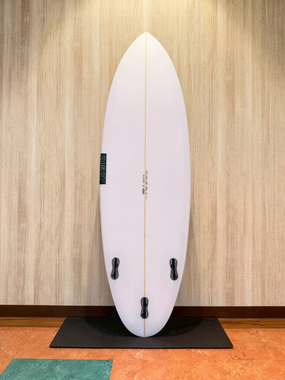 LINE_ALBUM_2026 フリーダム_260601_4 - SHOP NEWS | ムラサキスポーツオンラインストア 通販 White surfboard standing upright indoors against a wood wall on a black mat, with an orange center string and four fin boxes on the bottom half.